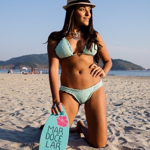 Pu&ograve; includere: Una donna in bikini di crochet blu chiaro e cappello di paglia &egrave; inginocchiata su una spiaggia di sabbia. Tiene in mano una tavola da surf blu e rosa con le parole "Mar Doce Lar" scritte su di essa.
