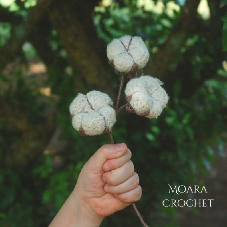May include: A hand holds a crocheted cotton boll stem with three white cotton bolls. The stem is brown and the bolls are white with brown accents. The background is out of focus and green. The text "MOARA CROCHET" is in the lower right corner of the image.