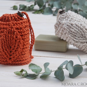 May include: Two crocheted soap savers, one in orange and one in beige, with a bar of soap and eucalyptus leaves on a white wooden surface. The text "MOARA CROCHET" is visible in the bottom right corner of the image.