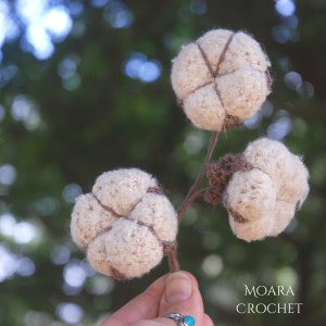 May include: A close-up of three crocheted cotton boll ornaments, each with a brown stem. The bolls are white and fluffy, with a brown crocheted detail in the center. The background is blurred and green.