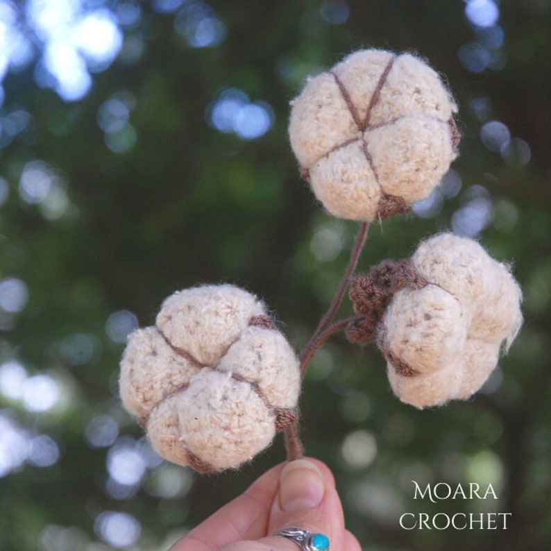 May include: A close-up of three crocheted cotton boll ornaments, each with a brown stem. The bolls are white and fluffy, with a brown crocheted detail in the center. The background is blurred and green.