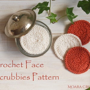 Puede incluir: Tres esponjas faciales de crochet, dos son naranjas y una es blanca. Las esponjas están en un frasco de vidrio con una tapa plateada. El texto "Crochet Face Scrubbies Pattern" está escrito en rojo debajo del frasco. El texto "MOARA CROCHET" está escrito en negro en la parte inferior de la imagen.