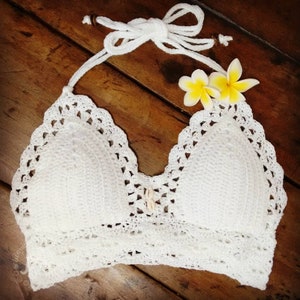 Pu&ograve; includere: Top bianco a halter in crochet con un bordo a scalare e un accento a conchiglia. Il top &egrave; su una superficie di legno con fiori gialli.