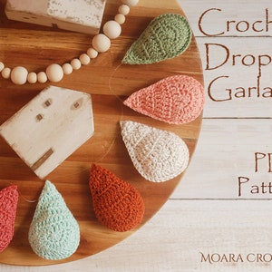May include: A crochet droplet garland pattern PDF. The image shows a wooden board with a garland of crocheted teardrop shapes in various colours, including green, pink, white, and orange. The text "Crochet Droplet Garland PDF Pattern" is written on a white background.