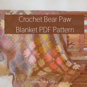 May include: A crocheted blanket with a bear paw pattern in shades of brown, pink, yellow, and peach. The blanket is draped over a wooden bench. Text on the blanket reads "Crochet Bear Paw Blanket PDF Pattern".