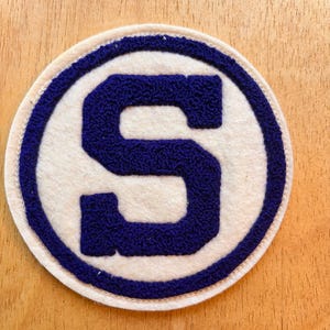 May include: A circular patch featuring a large, dark blue letter S in a textured, raised chenille embroidery style. The letter is centered on a cream-colored felt background, surrounded by a thick, dark blue circular border and a cream edge.