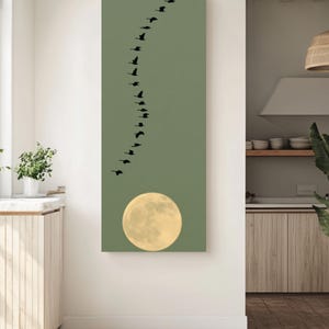 May include: A vertical rectangular wall art piece featuring a sage green background. A large, pale yellow full moon is positioned at the bottom center. Above the moon, a flock of black birds is arranged in a winding, S-shaped formation.