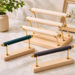 May include: A jewelry display stand set featuring a tiered rack with three beige velvet-covered bars and two separate single-bar stands in dark teal and dark gray velvet. Each stand has gold metal supports mounted on light-colored wooden bases.