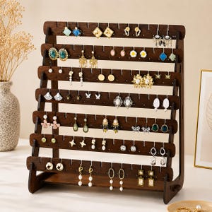 May include: A dark brown wooden earring display stand featuring seven horizontal tiers with evenly spaced notches along the top edges. The A-frame structure is designed to hold multiple pairs of dangling and stud earrings, showcasing a variety of jewelry styles arranged across the wooden slats.
