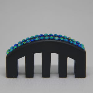 May include: A matte black hair comb featuring a curved top edge adorned with two parallel rows of small, iridescent blue and green crystals. The hair accessory has five sturdy, vertical teeth and a smooth, solid finish across the base structure.