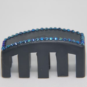 May include: A matte black hair claw clip featuring a rectangular top surface bordered by a single row of small, iridescent blue crystals that reflect light with hints of purple and green. The clip has four downward-facing prongs on each side.