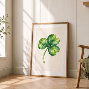 May include: A framed wall art print featuring a watercolour illustration of a single green three-leaf clover centred on a light cream background. The artwork is enclosed in a simple light-toned wooden frame and rests on a light-coloured floor.