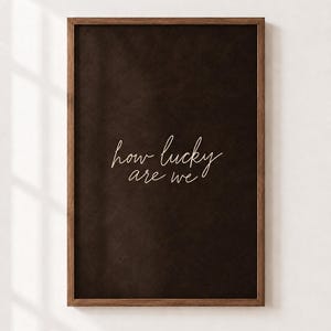 Pode incluir: Uma peça de arte de parede retangular com um fundo castanho escuro texturizado, inserida numa moldura de madeira fina. O texto "how lucky are we" está escrito numa fonte cursiva fina e clara, centrada na superfície escura.