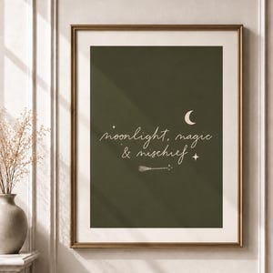 May include: A framed wall art print featuring a dark olive green background with the white text "moonlight, magic & mischief" in a cursive script. The design includes a crescent moon, small four-pointed stars, and a small broomstick icon.