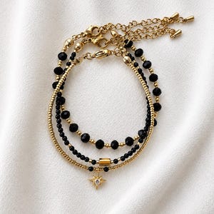 May include: A layered bracelet set featuring two strands of black faceted beads and gold-toned metallic beads. One strand includes a central gold-toned cylindrical bead, while the other features a gold-toned starburst charm with a central clear stone.