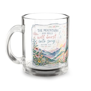 May include: A clear glass mug featuring a colorful illustration of mountains, hills, and wildflowers. The text reads: THE MOUNTAINS AND HILLS WILL burst into song BEFORE YOU ISAIAH 55:12. The design includes musical notes and a sunrise.