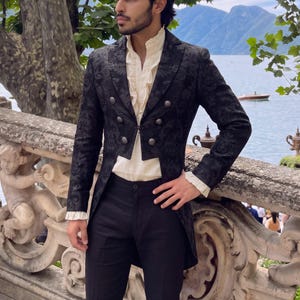 May include: A black double-breasted tailcoat featuring a tonal damask pattern and silver-toned buttons, worn over a cream-colored shirt with a ruffled front placket and ruffled cuffs, paired with solid black trousers.
