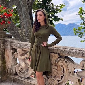 Könnte beinhalten: Ein olivgrünes Langarmkleid mit Kapuze, einer Schnürung im Korsett-Stil auf der Vorderseite und einem asymmetrischen Saum. Das Kleidungsstück hat ein eng anliegendes Oberteil, das in einen ausgestellten Rock übergeht und eine strukturierte Form.