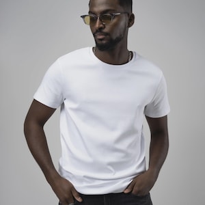 Può includere: Una t-shirt bianca a maniche corte con girocollo abbinata a jeans in denim grigio scuro. L'outfit è completato da una collana a catena argentata e da occhiali con montatura rettangolare, lenti colorate gialle e bordi grigio scuro.