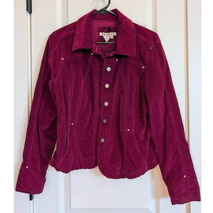 May include: A deep magenta corduroy jacket featuring a classic collar, long sleeves, and a front button-down closure. The garment includes vertical seam detailing, small metallic accents along the seams, and a label reading A.M.I. at the neck.
