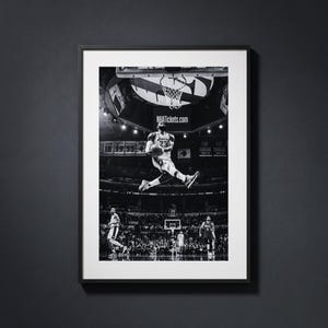 May include: A black and white framed photograph depicting a basketball player in mid-air performing a dunk during a game. The arena features a scoreboard with the text NBATickets.com and a crowd in the background. The frame is thin and black.