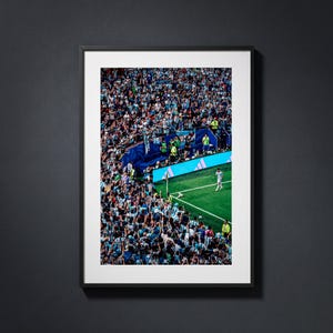 May include: A framed vertical photograph of a crowded soccer stadium featuring a large group of spectators in light blue and white jerseys, a green grass field, a corner flag, and a single player standing near the sideline by a blue wall.