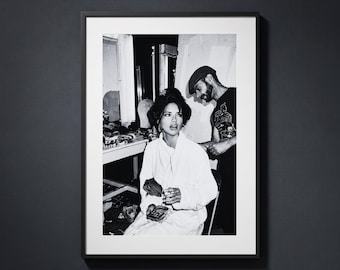 May include: A black and white framed photograph depicting a person in a white robe sitting and holding a small object, while another person wearing a cap and a dark t-shirt stands nearby in a room with various items on a table in the background.
