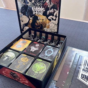 May include: The Unmatched Cobble & Fog board game box set features a vertical lid with gothic illustrations, an open base containing organized card decks with distinct graphic designs, and several miniature figures. A rulebook is included.