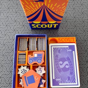 May include: A Scout card game set featuring an orange box with a blue circus tent graphic, a blue storage tray with stacks of gray tokens, a blue car token, star-shaped tokens labeled -20 and -5, and a deck of purple and white cards.