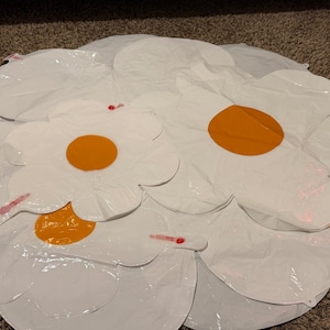 Puede incluir: Un conjunto de globos inflables blancos con forma de flor, cada uno con un centro circular naranja prominente. Estas decoraciones de plástico brillante y ligero están dispuestas en un grupo, mostrando sus contornos de pétalos y superficies lisas.