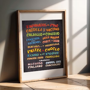 May include: A framed chalkboard-style print featuring a colorful menu of Chilean dishes. The text includes "Empanadas de Pino," "Cazuela de Vacuno," and other traditional food items. The frame is made of light-colored wood.