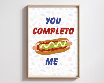 May include: A framed print featuring a cartoon hot dog with mustard and ketchup. The text "YOU COMPLETO ME" is written above and below the hot dog in blue and red. The background has a repeating pattern of small, colorful illustrations.