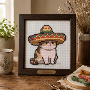 May include: A framed cross-stitch embroidery featuring a calico cat wearing a large, colorful sombrero with red, green, and yellow patterns. The dark wood frame includes a small brass plaque at the bottom with the text Aries Creations engraved.