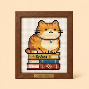 May include: A cross-stitch embroidery of an orange tabby cat sitting on a stack of three books, framed in dark wood. The cat has a white chest and a blue collar. The top book has a crown icon and the text "Foxy". A metal plaque reads "Aries Creations".