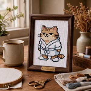 May include: A cross-stitch embroidery of a brown tabby cat wearing a white bathrobe and slippers, framed in a dark wood frame with a gold plaque reading Aries Creations. The design is centered on white Aida cloth with a pixelated aesthetic.