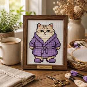 May include: A framed cross-stitch embroidery featuring a beige cat wearing a purple bathrobe and matching slippers. The wooden frame includes a small gold plaque at the bottom with the text Aries Creations printed in black lettering.