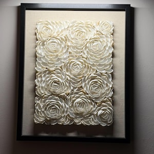 Peut inclure: Une œuvre d'art murale rectangulaire composée d'une collection de formes circulaires blanc cassé, superposées comme des pétales et disposées en grille. L'œuvre est encadrée par un cadre noir fin sur un fond en tissu beige clair.