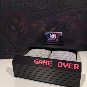 May include: A black storage unit for video game cartridges featuring a horizontal ribbed front panel with the text GAME OVER in pink pixelated letters. The top surface holds a small black display stand with a rectangular card labelled GAME OVER.