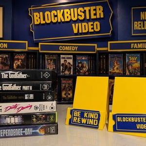 以下が含まれることがあります： The Godfather、Dirty Dancing、Men in Blackを含む6本のVHSテープの山と、Be Kind RewindおよびBlockbuster Videoと書かれた2つの黄色いディスプレイスタンド。背景にはジャンル名が記された映画の棚が並んでいる。