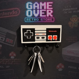 May include: A wall-mounted key holder shaped like a classic Nintendo NES controller with a white, black, and red color scheme. The controller features a directional pad, Select and Start buttons, and two red A and B buttons. Three black hooks extend from the bottom, holding a metal keyring with several keys attached.