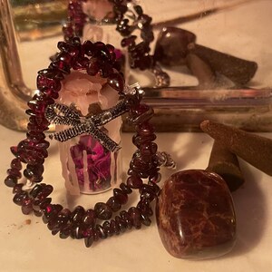 Könnte beinhalten: Ein kleines Glasgefäß gefüllt mit getrockneten violetten Blütenblättern, versiegelt mit Wachs und verziert mit einem silbernen Band. Das Glas ist von einer Kette aus dunkelroten Granat-Splitterperlen umgeben. Daneben liegt ein polierter Granatstein.