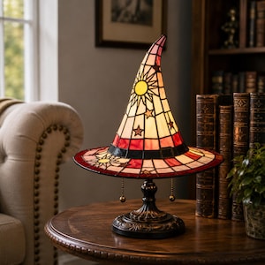 Peut inclure: Une lampe de table en vitrail en forme de chapeau de sorcière pointu. La lampe présente une palette de couleurs crème, rouge et noire avec des motifs de soleil et d'étoiles. Elle possède une base en métal sombre avec deux chaînettes.