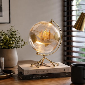 Op de afbeelding: Een decoratieve wereldbollamp met een heldere glazen bol met goudkleurige continenten en een miniatuur zeilschipmodel aan de binnenkant. De wereldbol wordt ondersteund door een goudkleurige metalen standaard met een driepootvoet en interne verlichting.