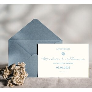 May include: A white rectangular save the date card with blue text reading "SAVE OUR DATE", "Michele & Thomas", "ARE GETTING MARRIED", and "07.04.2027" with a small blue shell icon, positioned in front of a dusty blue envelope and dried flowers.