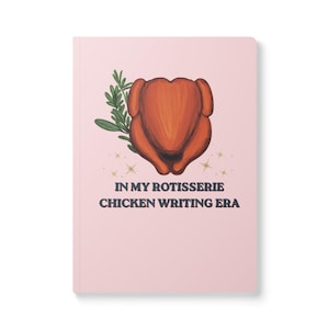 May include: A light pink notebook featuring a central illustration of a roasted rotisserie chicken with a sprig of green herbs. Below the chicken are small gold star icons and the text "IN MY ROTISSERIE CHICKEN WRITING ERA" in dark lettering.