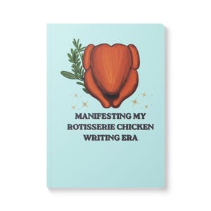 May include: A light blue notebook featuring a central illustration of a roasted rotisserie chicken with a sprig of green herbs. Below the chicken are small gold star icons and the text: MANIFESTING MY ROTISSERIE CHICKEN WRITING ERA.