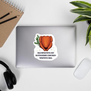 May include: A white sticker featuring a golden-brown roasted rotisserie chicken with a sprig of green rosemary, surrounded by small gold sparkles, and the black text: MANIFESTING MY ROTISSERIE CHICKEN WRITING ERA, centered on a silver laptop.