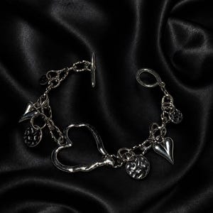 Könnte beinhalten: Ein silberfarbenes Bettelarmband mit Knebelverschluss, einem großen herzförmigen Glied, zwei massiven, gewölbten Herzanhängern und zwei kleinen, strukturierten, runden Münzanhängern, die an einer dicken Kette mit poliertem Metallfinish hängen.