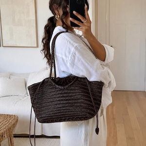 May include: A dark brown woven leather tote bag featuring a braided shoulder strap and a drawstring closure with thin leather ties. The bag has a textured, interlaced pattern throughout its exterior and a soft, gathered opening at the top.