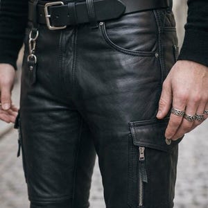 Peut inclure: Pantalon cargo en cuir noir avec une finition lisse, plusieurs poches à fermetures éclair argentées et une ceinture en cuir noir assortie avec une boucle en métal argenté. Le pantalon présente des coutures renforcées et un clip en métal.